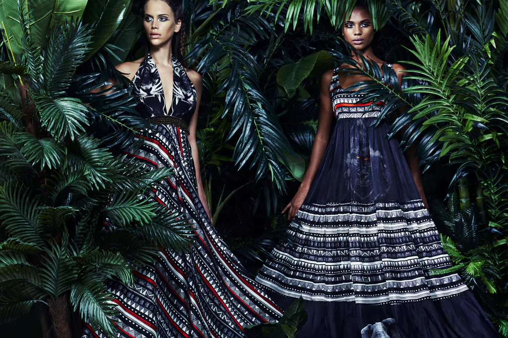 Editorial image of two models: one in tiger print bustier dress with African tribal prints; another in gypsy-style long dress with gorilla, elephant animal print, tribal motifs; green forest studio background
